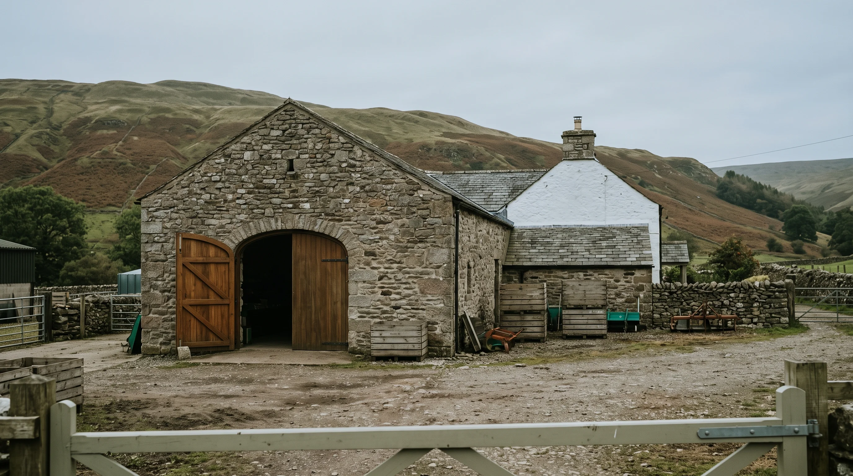 A farm shop in a converted stone barn with its door open onto the yard, cottage windows in the gable beyond and the fell behind under grey sky